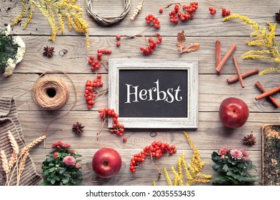 Text Herbst on blackboard means Autumn in German language. Orange Hokkaido pumpkins, rowan berry, apples, cinnamon and frame made from Fall decorations. Flat lay, top view on old, aged wooden table.