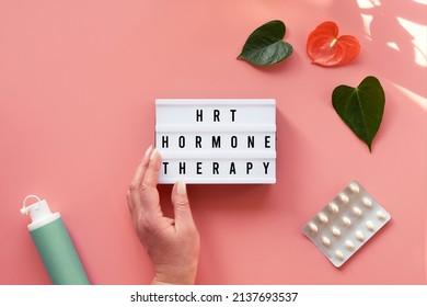 Text HRT Replacement Therapy On Light Box In Hand. Menopause, Hormone Therapy Concept. Estrogen Replacement Therapy Awareness Design. Pink Background With Exotic Leaves, Flowers, Pills, Estrogene Gel.