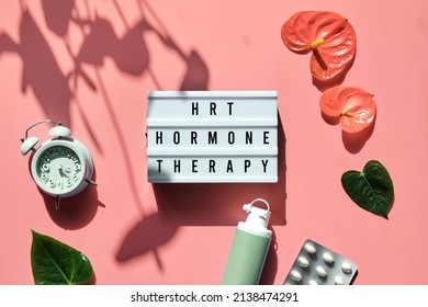 Text HRT Replacement Therapy On Light Box. Menopause, Hormone Therapy Concept. Oestrogen Replacement Therapy Awareness. Pink Background With Alarm Clock, Exotic Leaves, Pills, Estrogene Gel.