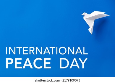 Text International Peace Day In White Letters On A Blue Background Next To A Flying White Paper Dove, Symbol Of Peace.