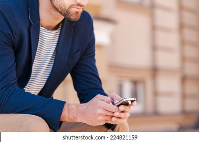 Text messaging. Cropped image of handsome young man in smart jacket typing something on mobile phone while sitting on the bench outdoors