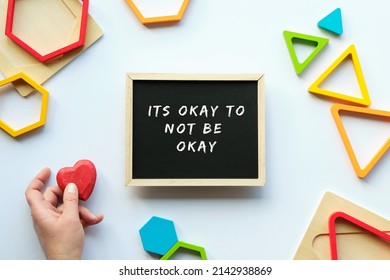 Text Its okey to not be okey on blackboard, chalk board. Nested wood triangles and hexagons in rainbow colors. Red wooden heart in human hand. Geometric education stacking puzzle pieces. Motivation
