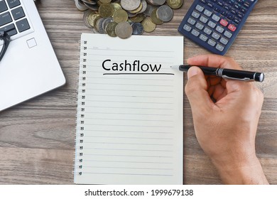 Text on cashflow written on the notebook with cropped had holding a pen.  Calculator, pile of coins, eyeglasses on the laptop keyboard.  Business and cashflow management concept