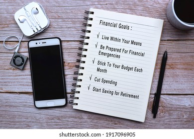 Text On Notepad With Cup Of Coffee, Smart Phone, Music Player And Pen On Wooden Desk - Financial Goals List