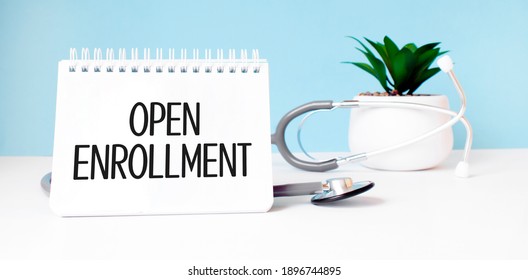 The text open enrollment is written on notepad near a stethoscope on a blue background. Medical concept