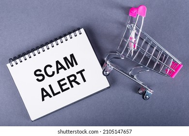 The Text SCAM ALERT Is Written On A Notebook, Next To A Trolley On Gray