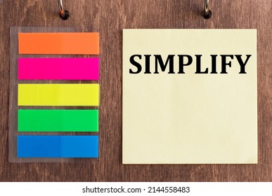 Text SIMPLIFY on a yellow card on a wooden background next to colored stickers for notes