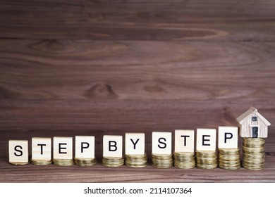Text STEP BY STEP made from wooden letter blocks and a miniature house placed on ascending coins stacks. Home buying process concept.