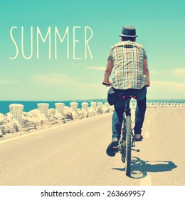 The Text Summer And A Back View Of An Active Young Caucasian Man Riding A Bike On A No Traffic Road Near The Sea, With A Filter Effect