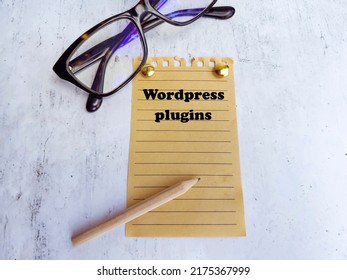 Text Wordpress Plugins written on notepaper with pencil and eyeglasses on gray  background.