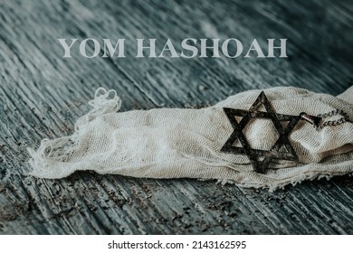 the text Yom Hashoah and an old and rusty pendant in the shape of the star of David on a piece of a ragged cloth, placed on a gray rustic wooden surface