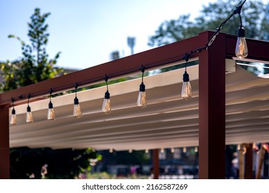 Textile Awning In The Backyard Wooden Frame Gazebo With A Garland Of Strings Of Retro Edison Light Bulbs Of Lamps Lighting Glowing With Warm Light On Sunny Summer Day Close-up, Nobody.