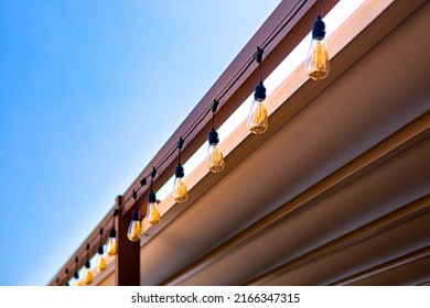 Textile Awning In Backyard Wooden Frame Gazebo With Garland Of Strings Of Retro Edison Light Bulbs Glow With Warm Light On The Patio, Summer Day Close-up Light Decor With Blue Sky, Nobody.