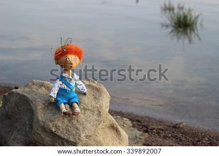 Textile handmade doll curious boy with dragonfly in a ginger hair in blue shorts on the stone near water