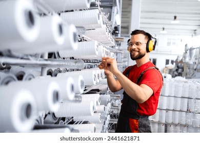 Textile industry. Factory worker with earmuffs standing by large industrial knitting machine and checking threads. - Powered by Shutterstock