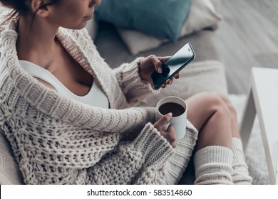Texting to boyfriend. Close-up of beautiful young woman holding cup and using smart phone while relaxing on couch at home