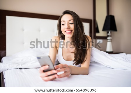 Texting to friend. Beautiful young woman smiling and holding smart phone while lying in the bed at home