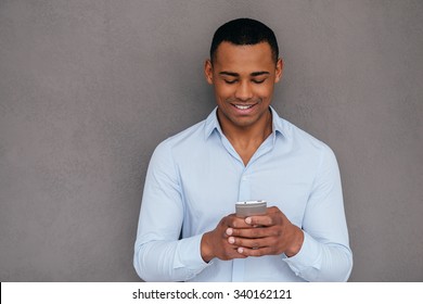 Texting To Friend. Confident Young African Man Looking At His Smart Phone And Smiling While Standing Against Grey Background