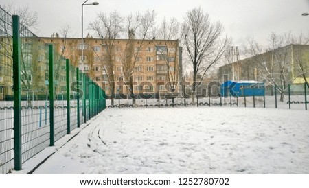 Texture and background of an old green metal fence or wicket in public places or sport fields against the background of residential buildings in the daytime in the winter time of the day. White snow.