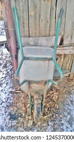 Texture And Background Of An Old Unicycle Metal Carriage Or Stroller For Household Needs, Attached To A Wooden Wall And The Last Autumn Leaves, A Shed, A Barn In The Winter Season During The Day.