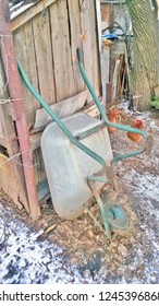 Texture And Background Of An Old Unicycle Metal Carriage Or Stroller For Household Needs, Attached To A Wooden Wall And The Last Autumn Leaves, A Shed, A Barn In The Winter Season During The Day.