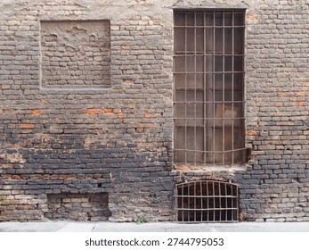 A textured, aged brick wall features a tall barred window, a small barred basement window, and a square opening sealed with bricks and plaster