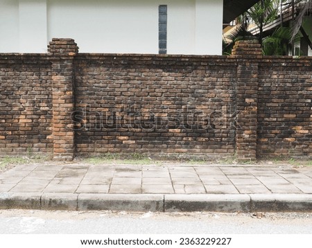 Textured and faded red brick wall with grass and moss standing on grey pavement and kerb
