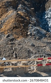 Textured landscape of construction zone featuring large mounds of excavated earth, mixed rubble, deep trenches, and temporary wooden safety barrier in foreground.