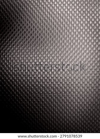 textured surface with a repeating geometric pattern. The surface features a dark, metallic-looking grid or mesh structure with a gradient of lighting, fading from deep shadows on the left to brighter highlights on the right.