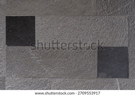 Textured wall surface composed of large grey rectangular natural stone tiles with rough surfaces and two smooth black inset stones