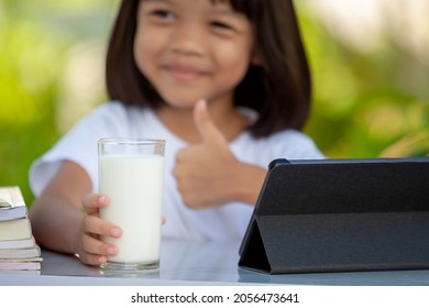 Thai Asian Kid Girl, Age 4 To 6 Years Old, Looks Cute And Bright. Growing Up, Holding A Glass Of Milk And A Tablet On The Table. Background Is Green. Selective Focus On Hands And A Glass Of Milk.