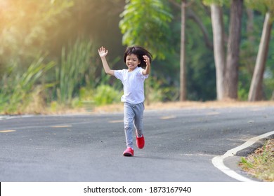 Thai Asian Kid Girl Aged 4 To 6 Years With Short Hair, Cute Face, And White Shirts. Running For Exercise And Playing On The Road. For Good Health, The Background Is A Refreshing Green Tree.