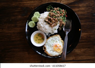 Thai food signature,Pad Kra Pao Khai Dow.Grill pork fry with herbs and natural in gredients.In black dish with spoon, fork, cup of suace place on dark brown table.