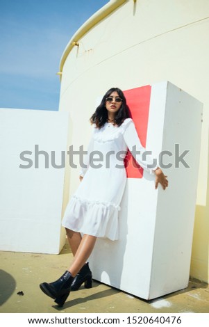 Thai girl in white dress wearing sunglasses and black boots leaning at white block on roof top.
