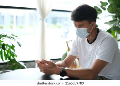 Thai Man Wearing The Medical Mask Using His Mobile Phone And Reading News About The Situation With Coronavirus Pandemic In The Coffee Shop. Daily Life After Covid-19. New Normal Lifestyle Concept.