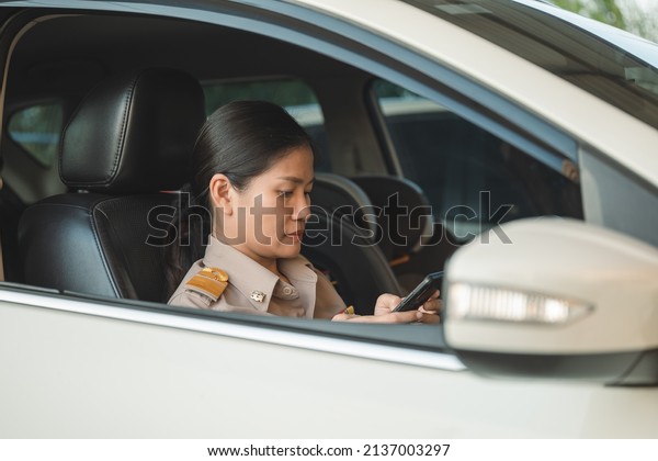 Thai teachers in official\
uniform making a serious expression while looking at her phone in a\
car.