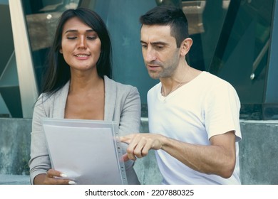 Thai Woman And Caucasian Man Reading Report Outside Glass Building. Office Teamwork Couple Discussing Project During Break At Work. Multi Ethnic Colleagues, Business Coworkers Concepts