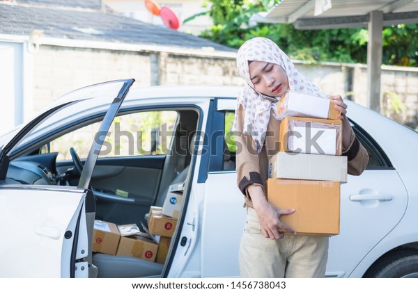Thai young muslim female messenger or delivery\
person talking on smartphone and holding many cardboard package\
boxes near the car.