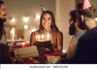 Thank You For The Surprise, Guys. Portrait Of Happy Young Woman In Her Twenties Surrounded By Friends, Holding Her Birthday Cake With Burning Candles And Looking At Camera During Party At Home Or Cafe