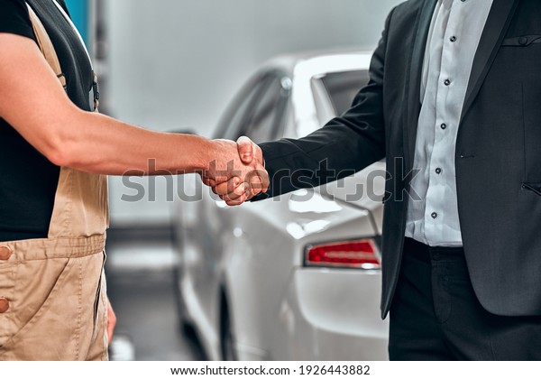 Thank you for your trust. Cropped shot of a\
mechanic shaking hands with the car service client repaired car on\
the background. Close up\
view.
