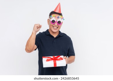 A thankful middle-aged Asian man wearing a party hat and novelty glasses celebrating, holding a gift at a birthday celebration, isolated on white. - Powered by Shutterstock