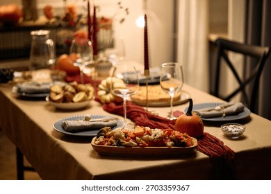 Thanksgiving dinner table set with assorted dishes, empty wine glasses, pumpkins, candles, serving platters and napkins arranged for gathering among friends, no people visible