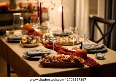 Thanksgiving dinner table set with assorted dishes, empty wine glasses, pumpkins, candles, serving platters and napkins arranged for gathering among friends, no people visible