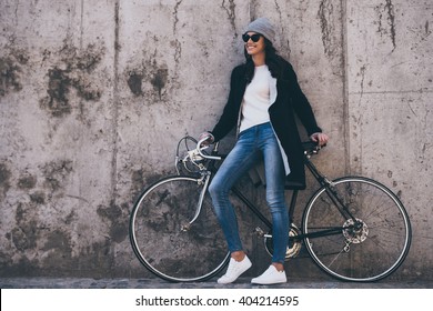 That was great ride! Beautiful young woman in sunglasses holding hands on her bicycle and looking away with smile while standing against concrete wall outdoors