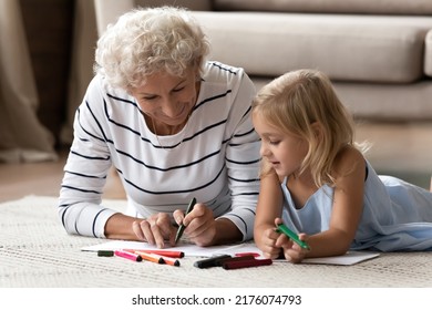 That is how to paint it. Caring mature granny teaching interested granddaughter preschooler to draw sketches on paper sheets, elderly nanny and little girl lying on warm floor indoor coloring pictures - Powered by Shutterstock