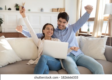 That Is It! Happy Laughing Young Married Couple Millennial Family Husband And Wife Sitting On Sofa At Home With Laptop Feeling Excited Joyful Receiving Good News, Purchasing Tour On Vacation Of Dream