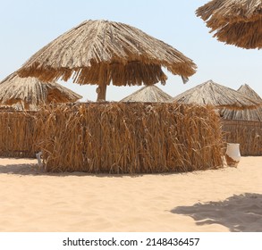 Thatched Sunshades And Furniture For Tourists, On A Yellow Sand Beach. Egyptian Hotel Beach. Concept Of Sea Vacations And Relaxation. Empty Sunbeds, Minimalistic Photo In Beige Tones.