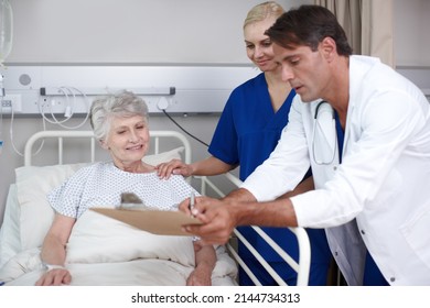 Thats Good News. A Doctor And His Young Nurse Showing Her Where To Sign The Medical Report While Standing Over Her Bed.