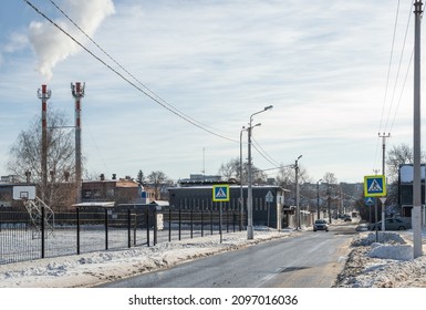 Thaw On Sunny Winter Day In Small Town, Narrow Street With Road And Poles,  Fence And Industrial Pipes In The Distance