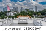 The Thean Hou Temple terrace. Ornaments on the white carved stone railings. The roof of the pagoda on a background of green tree. Skyscrapers, car parking in the distance. Blue sky, clouds. Malaysia.